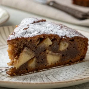 A close-up of a slice of chocolate apple cake, showing the moist texture and apple chunks inside, lightly dusted with powdered sugar.