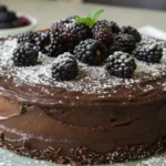 A close-up of a chocolate blackberry cake topped with fresh blackberries, powdered sugar, and a sprig of mint, served on a plate.
