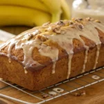 Freshly baked banana bread topped with a smooth glaze and sprinkled with chopped nuts, resting on a cooling rack with bananas in the background.