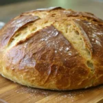 A close-up of a freshly baked round loaf of Miche bread with a golden-brown crust and visible cracks, resting on a wooden cutting board.