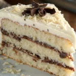 A close-up of a slice of White German Chocolate Cake showing moist layers with chocolate frosting, topped with shredded coconut and chocolate curls.