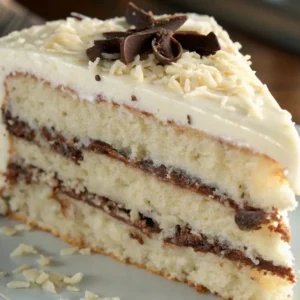 A close-up of a slice of White German Chocolate Cake showing moist layers with chocolate frosting, topped with shredded coconut and chocolate curls.