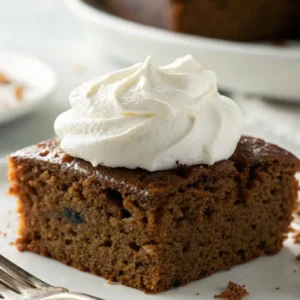 Close-up of a moist gingerbread cake square topped with a swirl of whipped cream on a white plate.