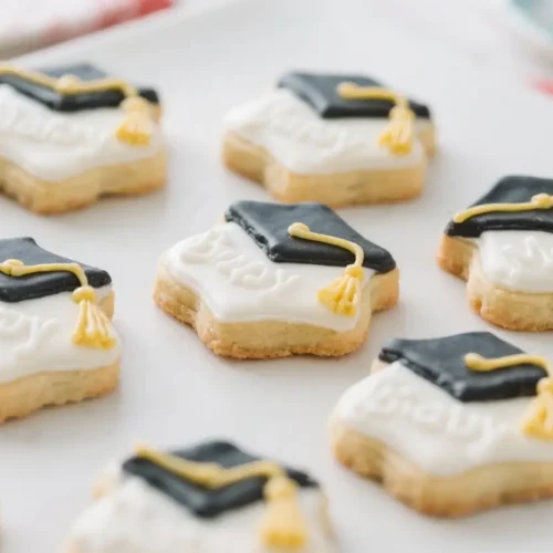 Decorated graduation cookies shaped like caps, iced in white and black with yellow tassel details, arranged on a tray.