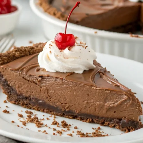 Slice of chocolate silk pie with creamy chocolate filling on a plate topped with whipped cream and a cherry, with the remaining pie in the background.