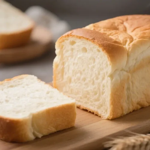 Fluffy Shokupan Japanese White Bread Recipe - Soft, Cloud-Like Loaf 2 Close-up of freshly baked Shokupan Japanese white bread loaf sliced open on a wooden board, revealing ultra-fluffy, pillowy interior with thin golden crust, accompanied by smaller loaves and wheat stalks on a rustic gray surface.