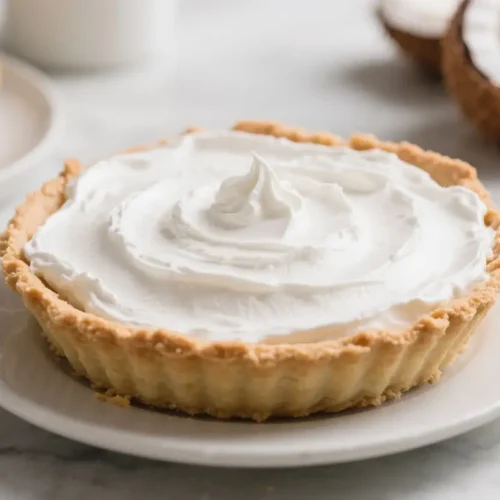 Coconut cream pie with a flaky crust and a thick layer of whipped cream topping on a white plate.