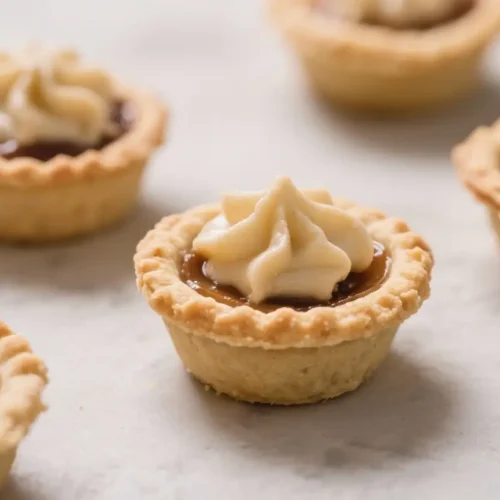 Mini pies in flaky pastry crusts filled with caramel and topped with a swirl of whipped cream on a light surface.