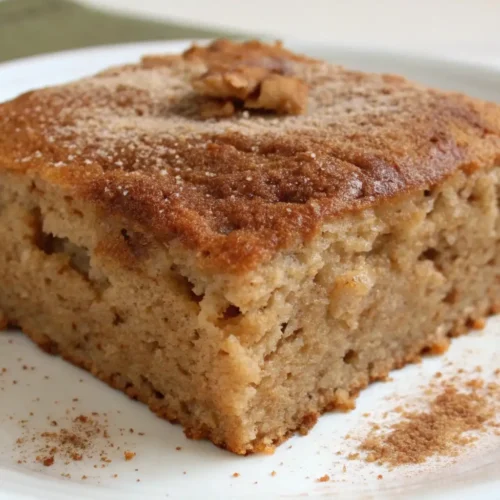 Moist applesauce snack cake slice on a white plate dusted with cinnamon and topped with a walnut.