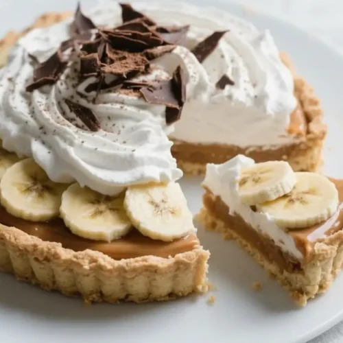 Slice of banoffee pie with banana slices, caramel toffee filling, and whipped cream topped with chocolate shavings in a buttery tart crust.