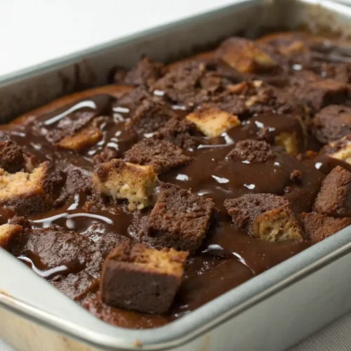 A rectangular metal baking pan filled with chocolate bread pudding, featuring toasted cubes of crusty bread soaked in a deep cocoa custard and topped with a glossy, melted chocolate ganache.