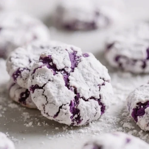 A close-up view of several deep purple ube crinkle cookies coated with white powdered sugar, scattered with extra sugar, sitting on a light-colored surface with a blurred background.