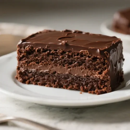 A close-up photograph of a rectangular slice of rich chocolate cake with a dark, textured crumb, filled with a central layer of chocolate cream and topped with a glossy, smooth chocolate glaze, sitting on a white plate with a silver fork nearby and chocolate pieces in the background.