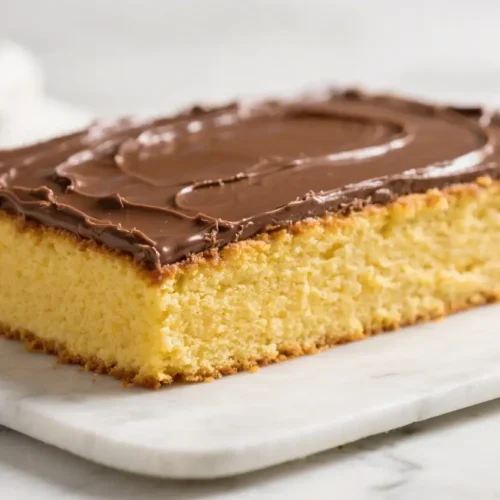 Moist yellow sheet cake topped with smooth chocolate frosting on a white marble board, close up showing soft fluffy crumb and glossy icing