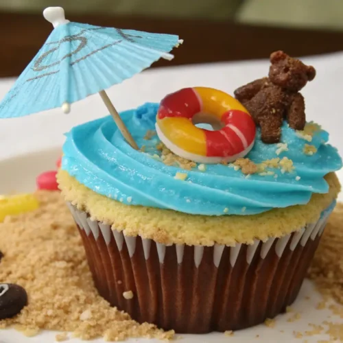 Beach-themed cupcake with blue frosting, mini umbrella, teddy bear, and candy life preserver on a sandy crumb base