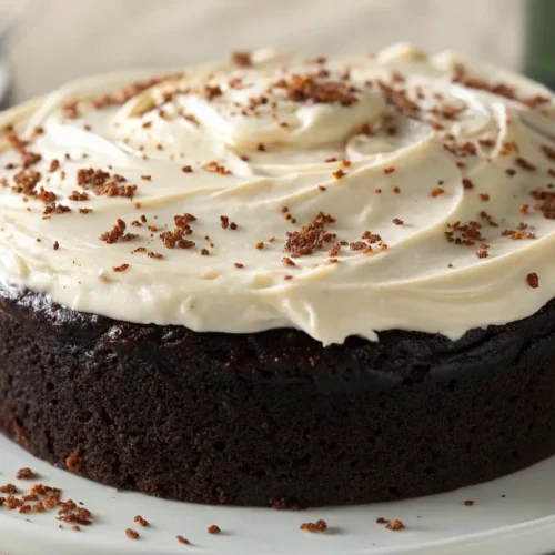 Moist chocolate Guinness cake topped with creamy white frosting and chocolate crumbs, served on a white plate with a blurred background.
