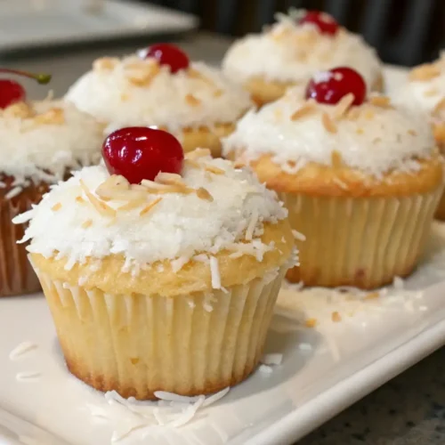 Fluffy Coconut Cupcakes With Smooth Frosting - Light, Tender, and Irresistible 2 Coconut cupcakes topped with white frosting, shredded coconut, toasted flakes, and red cherries arranged on a serving plate