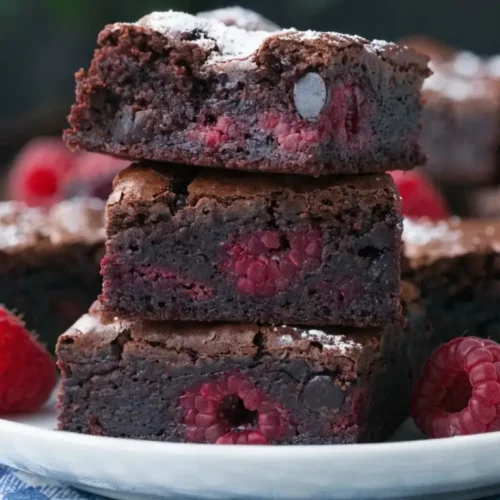 Stack of fudgy raspberry brownies on a plate with visible fresh raspberries inside, dusted with powdered sugar and surrounded by whole berries, showing a rich dense chocolate texture.