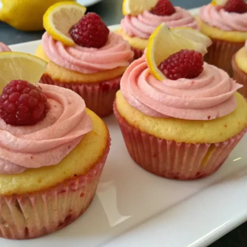 Lemon raspberry cupcakes with pink raspberry frosting, topped with fresh raspberries and lemon slices on a white plate.