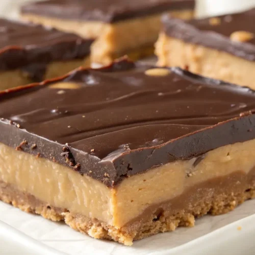 Buckeye fudge squares with creamy peanut butter filling, rich chocolate topping, and a crumbly cookie-style base on a white plate.