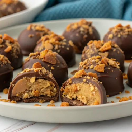 Chocolate-covered Butterfinger truffles arranged on a light gray plate, topped with crushed Butterfinger candy pieces. Two truffles are cut in half to reveal a creamy peanut butter filling with crunchy candy bits inside.