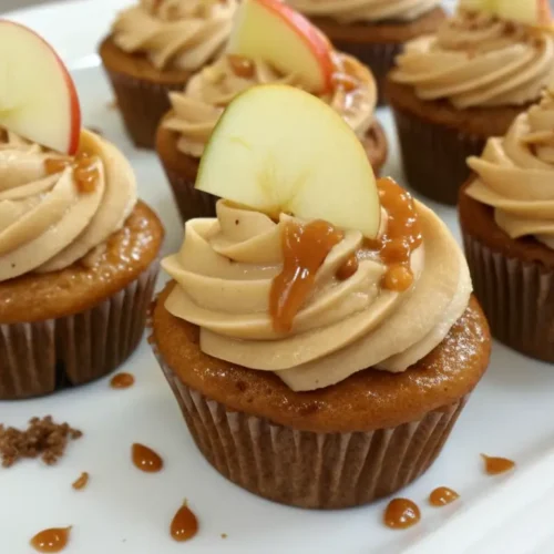 Caramel apple cupcakes topped with swirled caramel frosting, caramel drizzle, and fresh apple slices, arranged on a white serving plate.