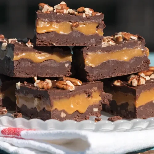 Caramel Pecan Turtle Fudge squares stacked on a white plate, showing rich chocolate layers, gooey caramel filling, and chopped pecans on top.