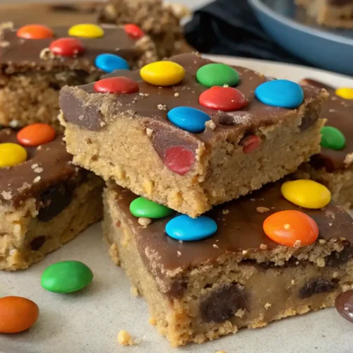 No Bake M&M Cookie Dough Bars topped with chocolate and colorful M&M candies, stacked on a plate with extra candies scattered around.