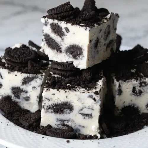Oreo fudge squares with a creamy white base and crushed chocolate sandwich cookies, stacked on a white plate with cookie crumbs scattered around.