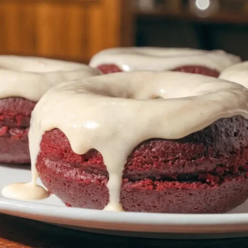 Red velvet cake donuts topped with thick cream cheese glaze, served on a white plate with glossy icing dripping down the sides.